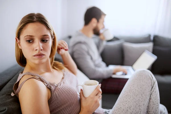 年轻迷人的金发姑娘早上坐在沙发上喝咖啡 而她的丈夫则用笔记本电脑对她不屑一顾 — 图库照片