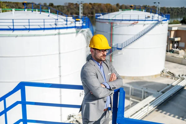 Young rich businessman in suit with helmet on head standing on height with arms crossed and looking around. Refinery exterior.