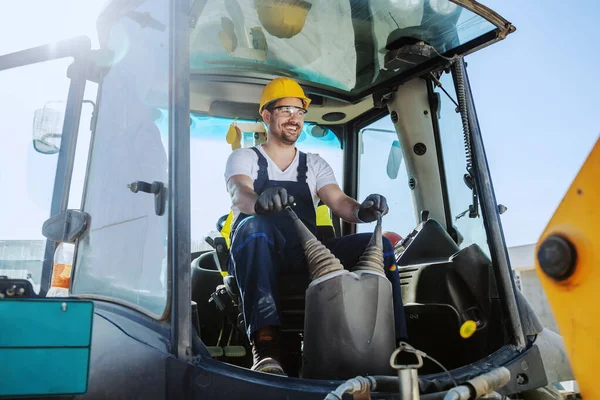 Smiling handsome caucasian worker in overall and with helmet on head driving excavator.