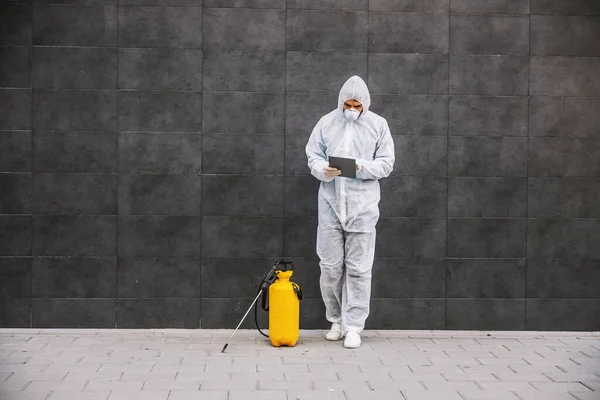 人はウイルスの保護のスーツおよびマスクを見 タブレットでタイプし 噴霧器とのコロナウイルスの建物を消毒する 感染予防と流行の制御 世界大流行 — ストック写真