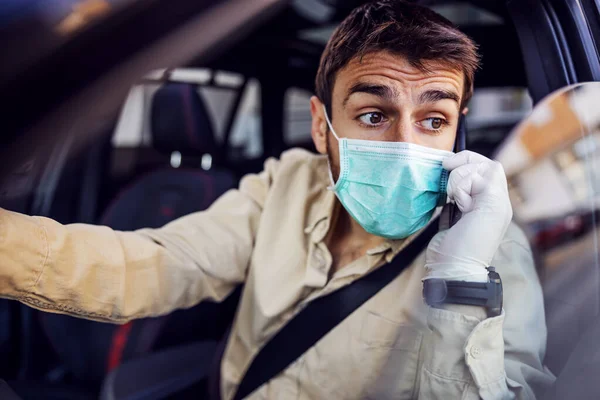 Man with protective mask and gloves driving a car talking on mobile phone smartphone. Infection prevention and control of epidemic. World pandemic. Stay safe.