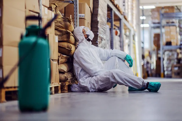 防護服を着た疲れた男と倉庫の床に座っているマスクと食品の完全な倉庫を消毒から休息 — ストック写真