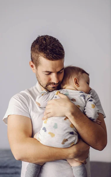Portrait of attractive unshaven young father holding his only beloved son and putting him to sleep. It\'s noon nap time.