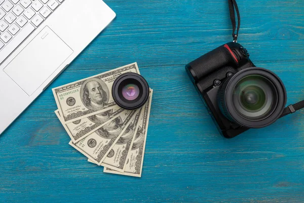 Earn money in internet DSLR camera, dollars, laptop, on blue wooden background, top view