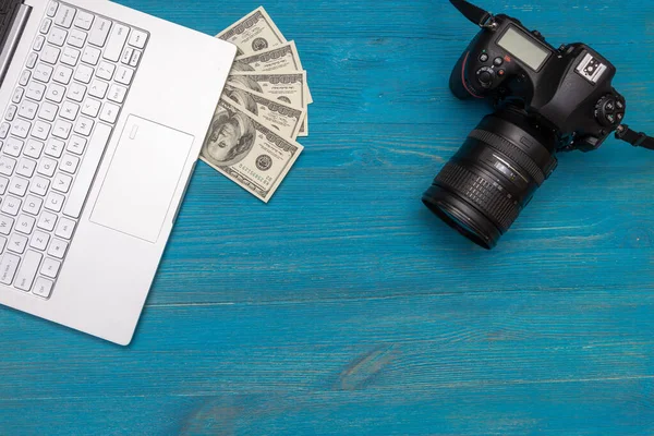 Earn money in internet DSLR camera, dollars, laptop, on blue wooden background, top view