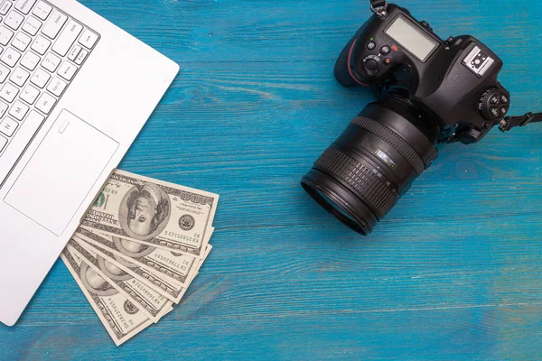 Earn money in internet DSLR camera, dollars, laptop, on blue wooden background, top view