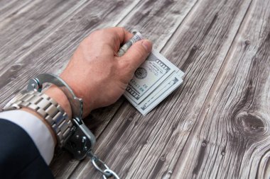 A man's hand in handcuffs and US dollars banknotes on wooden table. Corruption or economic crime concept.