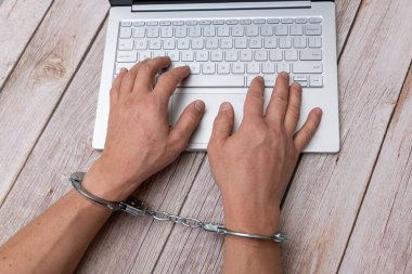 A man's hands in handcuffs and laptop on wooden table. Piracy, internet hacking and punishment for cybercrime concept.