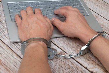 A man's hands in handcuffs and laptop on wooden table. Piracy, internet hacking and punishment for cybercrime concept.