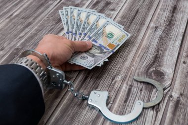 A man's hand in handcuffs and US dollars banknotes on wooden table. Corruption or economic crime concept.
