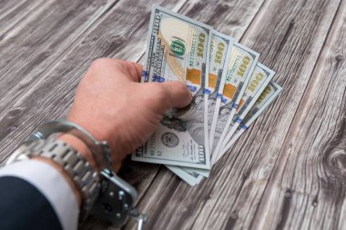A man's hand in handcuffs and US dollars banknotes on wooden table. Corruption or economic crime concept.