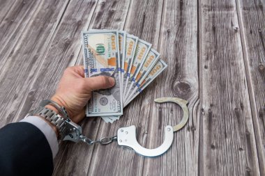 A man's hand in handcuffs and US dollars banknotes on wooden table. Corruption or economic crime concept.