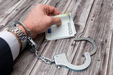 A man's hand in handcuffs and US dollars banknotes on wooden table. Corruption or economic crime concept.
