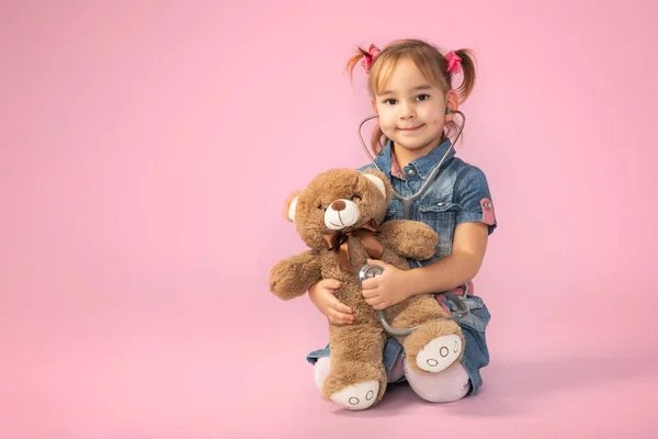 Happy Child Girl Doctor Examining Her Dear Toy Bear on Pink Background