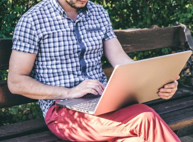 Genç adamın şehir parkında oturan ve onun laptopunda yazma gömlek giydiğini