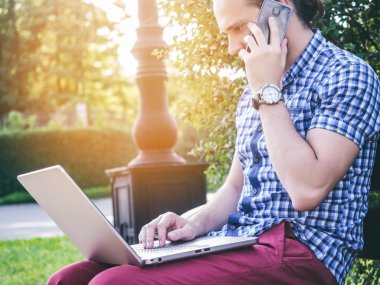 Yanında dizüstü bilgisayar ile smartphone dizlerinin üzerine konuşan genç adam