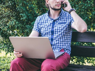 Yanında dizüstü bilgisayar ile smartphone dizlerinin üzerine konuşan genç adam