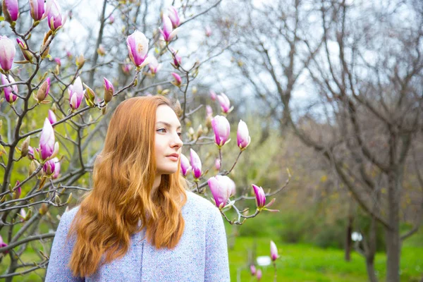 Bela mulher ruiva jovem de pé perto de magnólia florescente — Fotografia de Stock