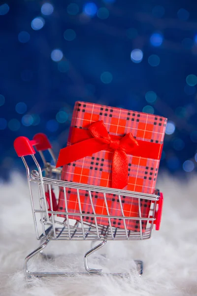 Scatola regalo rossa nel piccolo carrello del supermercato su blu bokeh backgrou — Foto Stock