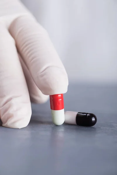 Closeup hand in medical latex glove holding two different capsules on gray table — Stock Photo, Image