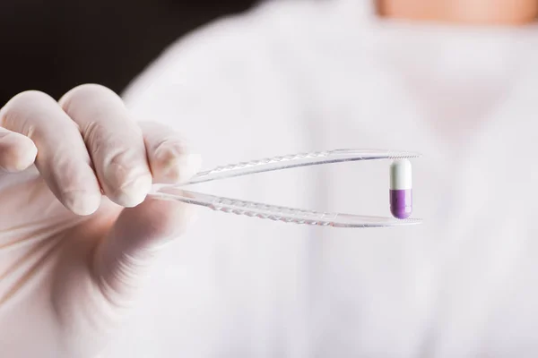 Hand in latex glove holding tweezers with violet and white capsule. Pharmaceutical research. — Stock Photo, Image