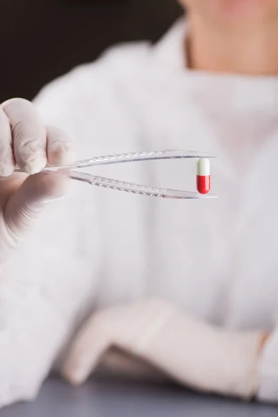 Hand in latex glove holding tweezers with red and white capsule. Pharmaceutical research. — Stock Photo, Image