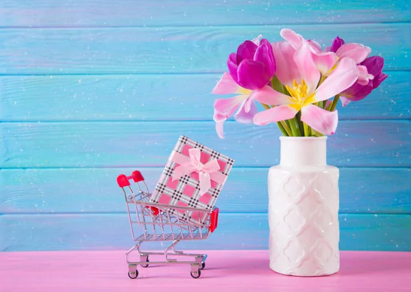 Petit chariot supermarché avec boîte cadeau et bouquet tendre de belles tulipes roses dans un vase sur fond de bois bleu et rose — Photo