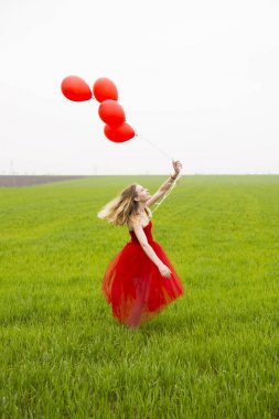 Kırmızı elbiseli sarışın saçlı güzel genç kız balon tutar ve yeşil yaz alanında fırtınası