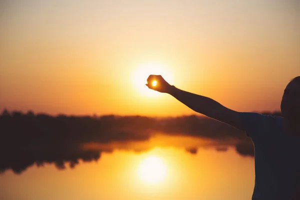 Силуэт улыбающегося молодого человека, держащего руку на солнце на закате у озера Стоковое Изображение