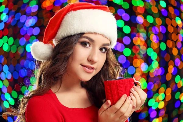 Young woman in santa hat with cup of tea on lights background — Stock Photo, Image