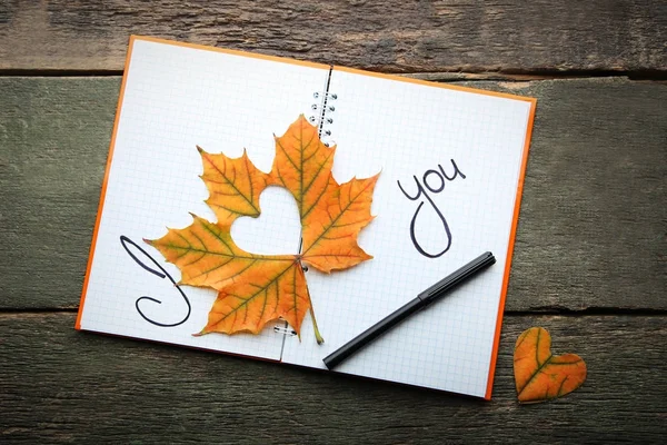 Dry maple leaf with heart and notebook on grey wooden table