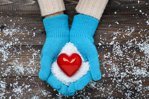 Mãos em luvas de malha segurando coração vermelho e neve em madeira ta — Fotografia de Stock