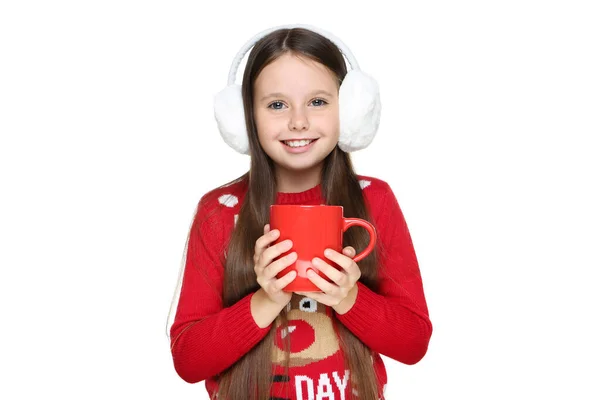 Egy kislány karácsonyi pulóverben és fülvédőben, kezében egy csésze tea fehér háttérrel. — Stock Fotó