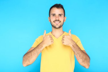 Mavi arka planda baş parmak gösteren genç sakallı adamın portresi.
