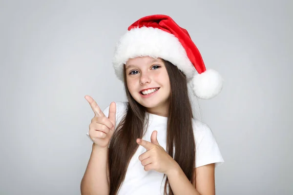Noel şapkalı küçük kız gri arka planda parmaklarıyla işaret ediyor. — Stok fotoğraf