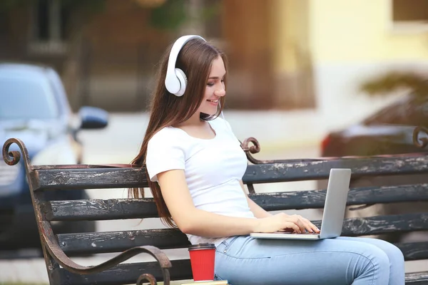 Молода дівчина з навушниками та ноутбуком сидить на лавці — стокове фото