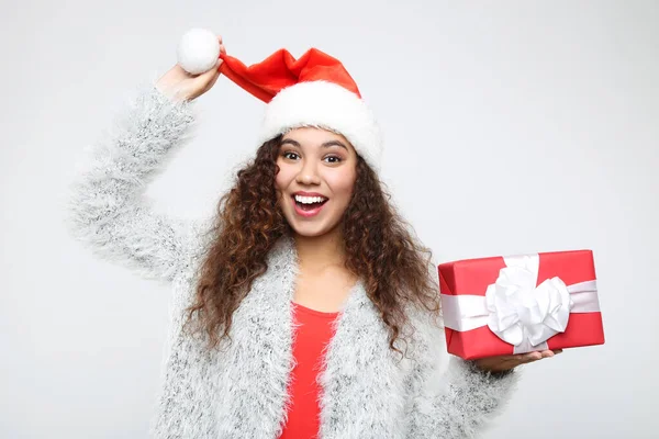 Hermosa mujer americana en santa hat sosteniendo caja de regalo sobre fondo gris —  Fotos de Stock