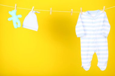 Sarı arka planda yumuşak oyuncaklı bebek kıyafetleri asılı.