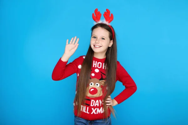 Κοριτσάκι σε χριστουγεννιάτικο πουλόβερ και κόκκινα κέρατα κυματίζει με το χέρι σε μπλε φόντο — Φωτογραφία Αρχείου