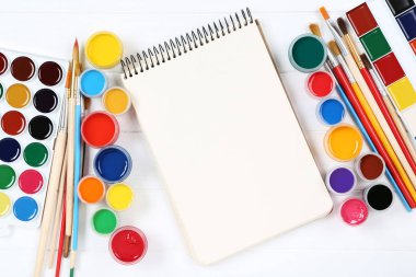 Fırçalarla renkli guaj boyaları ve beyaz ahşap masada boş kağıtlar.