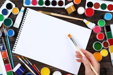 Kahverengi ahşap masa üzerinde renkli gouache boyaları ve fırçalar olan boş bir kağıt yaprağı.