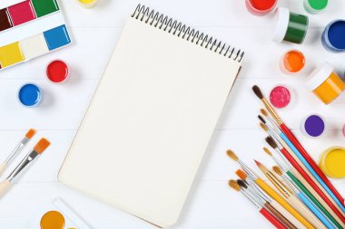 Fırçalarla renkli guaj boyaları ve beyaz ahşap masada boş kağıtlar.