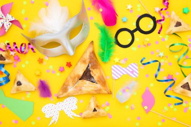 Purim tatil kompozisyonu. Sarı arka planda parti malzemeleri olan kurabiyeler.