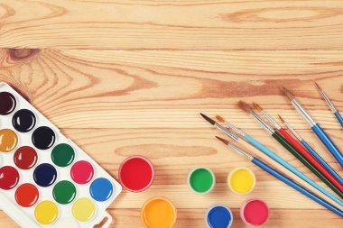 Kahverengi ahşap masada renkli guaj boyaları ve fırçalar.