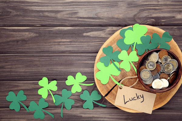 День Святого Патрика. Бумажные листья клевера с монетами в миске и слово Счастливчик на деревянном столе
