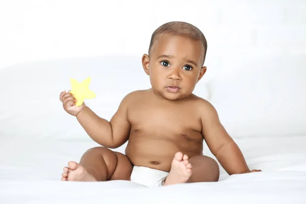 アメリカの赤ちゃん女の子座っていますホワイトベッドとホールディング黄色のおもちゃ — ストック写真
