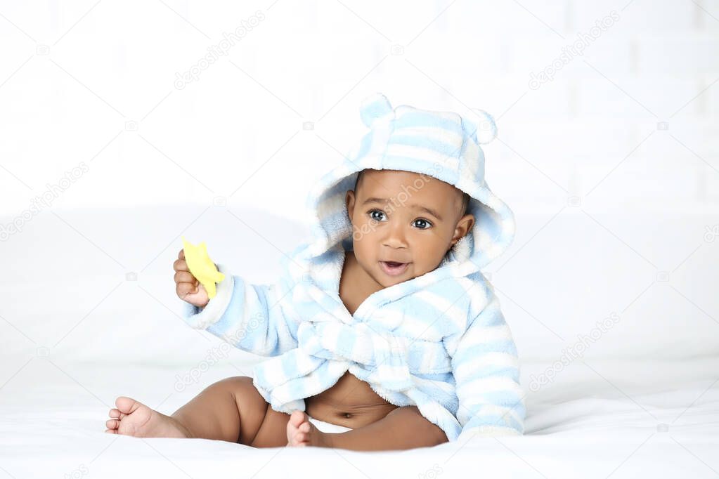 American baby girl in bathrobe sitting on bed and holding toy