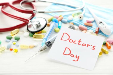 Doktor Günü 'nü steteskopla, renkli haplarla ve beyaz ahşap masada şırıngayla yaz.