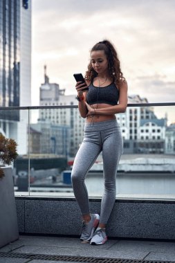 Spor elbiseli genç bir kadın şehrin arka planında telefonuyla eğitim sonuçlarını yayınlıyor.