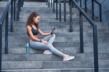 Spor elbiseli genç bir bayan stadyumun merdivenlerinde oturuyor ve elinde telefonla antrenman sonuçlarını yayınlıyor.